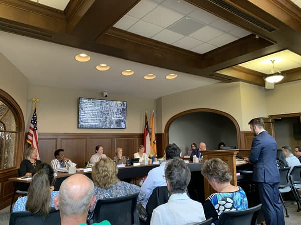 Man standing at podium in front of counsel members and audience.