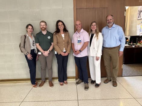 Six people posing for pic outside room.